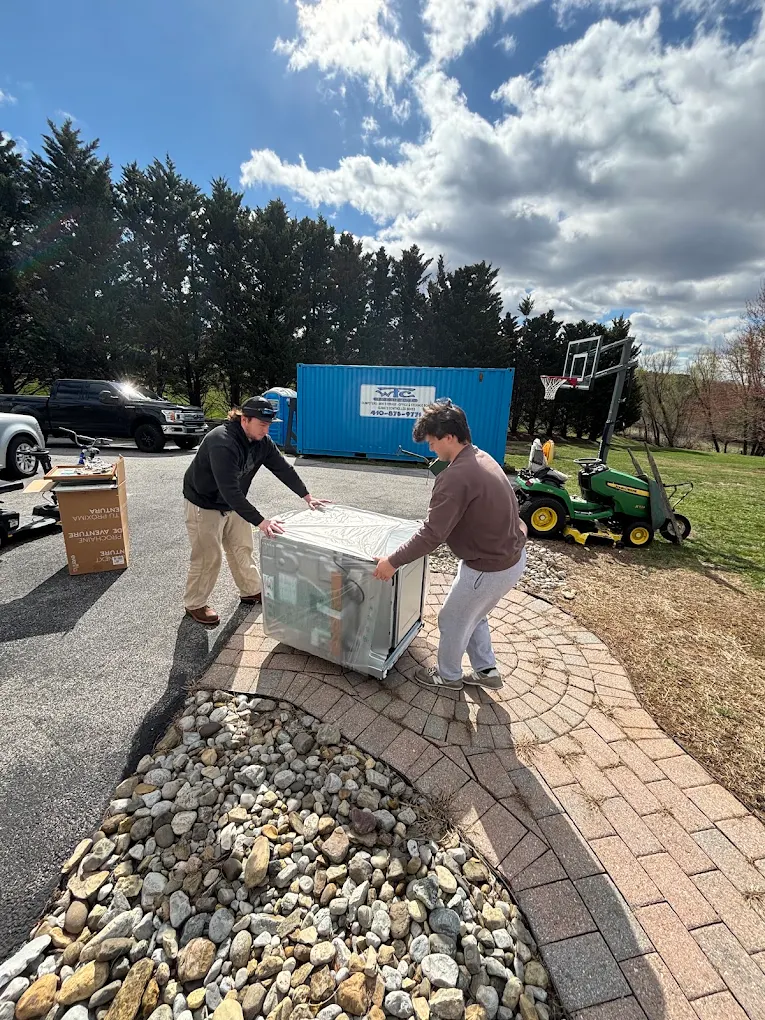 Two movers carrying a wrapped appliance across a paver driveway
