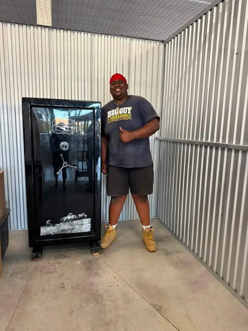 Aaron in a Big Guy Moving shirt beside a black gun safe inside a storage unit