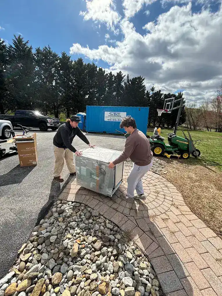 Two movers carrying a wrapped appliance across a paver driveway